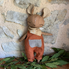 Charger l'image dans la galerie, Handmade brown linen mouse spirit guardian doll standing on fresh green bay laurel sprigs against a stone background, dressed in burnt orange overalls with a blue and white striped pocket and a beige scarf adorned with sky blue stars