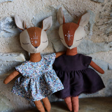 Charger l'image dans la galerie, Handmade linen fawn and fox dolls dressed in matching outfits—one in a floral dress, the other in deep plum—leaning against a stone wall in an embrace, with a sprig of delicate star-shaped flowers in the background.