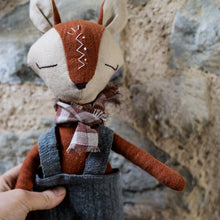 Charger l'image dans la galerie, Handmade linen fox spirit guardian doll wearing rustic overalls and a plaid scarf, held against a stone wall, featuring a hand-embroidered zig-zag tribal-inspired symbol on its forehead